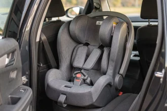 Siège auto bébé installé dans une voiture familiale, vue 3/4 depuis la portière ouverte montrant les harnais et protections latérales