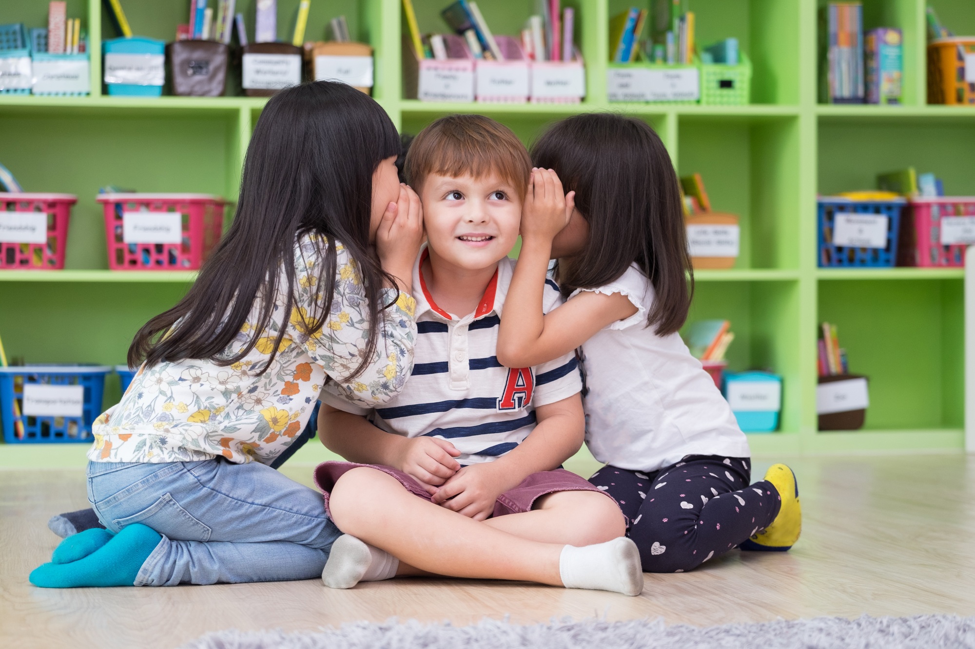 Two girl kids whisper secret at ear of boy in library at kindergarten